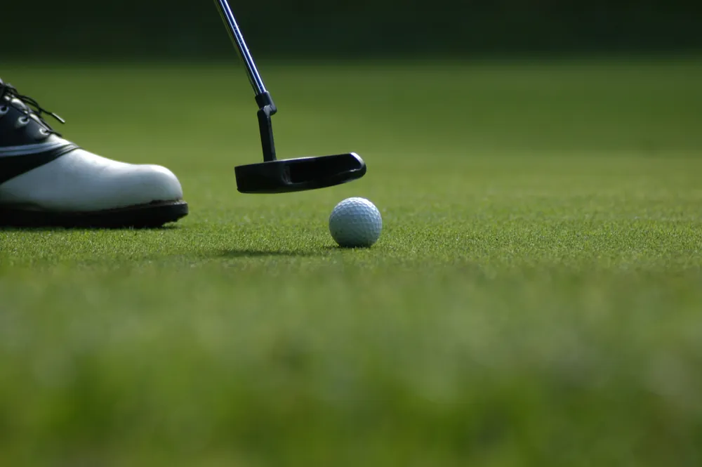 Putter head beside a white golf ball on a putting green, with a golfer’s shoe at the left.
